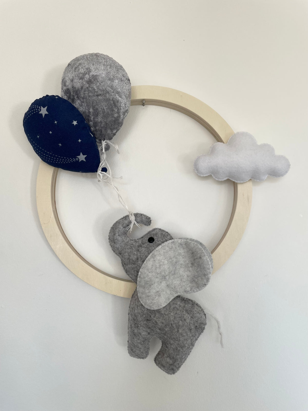 Couronne murale bébé faite main de 22cm en bois naturel avec un éléphant en feutrine, un ballon gris en velours, un ballon bleu marine étoilé et un nuage blanc.