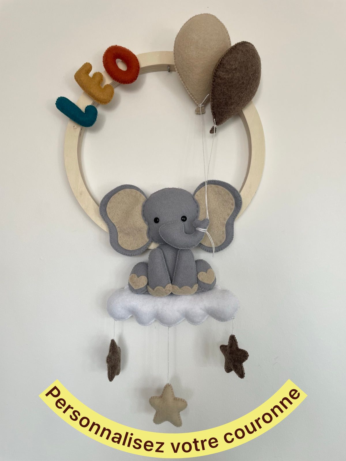 Décoration murale personnalisée pour chambre de bébé représentant une couronne en bois avec un éléphant installé sur un nuage, des ballons et des étoiles suspendues. Le prénom et les couleurs sont personnalisables. Création artisanale 