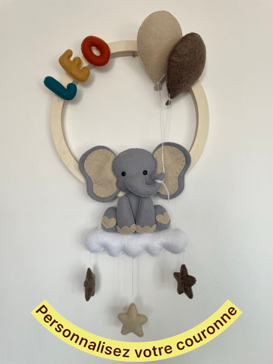 Décoration murale personnalisée pour chambre de bébé représentant une couronne en bois avec un éléphant installé sur un nuage, des ballons et des étoiles suspendues. Le prénom et les couleurs sont personnalisables. Création artisanale 
